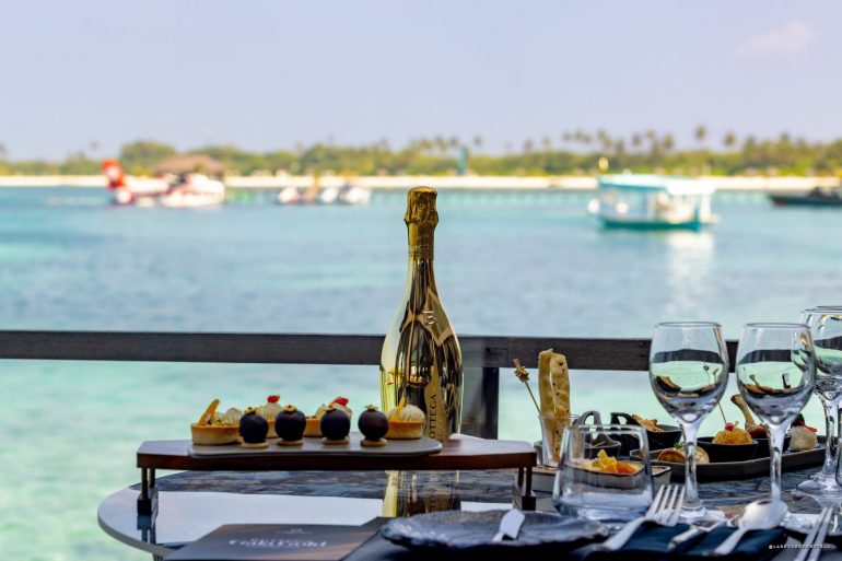 Champagne bottle centerpiece on a table with desserts and wine glasses, overlooking turquoise sea and boats in the distance.