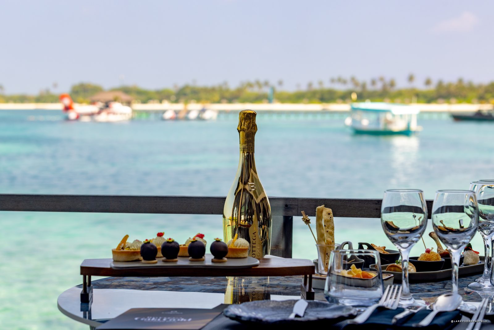 Champagne bottle centerpiece on a table with desserts and wine glasses, overlooking turquoise sea and boats in the distance.