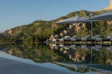 Row of white pool loungers with folded towels under large umbrellas by a calm reflecting pool, with green hills in the background, and a clear blue sky.