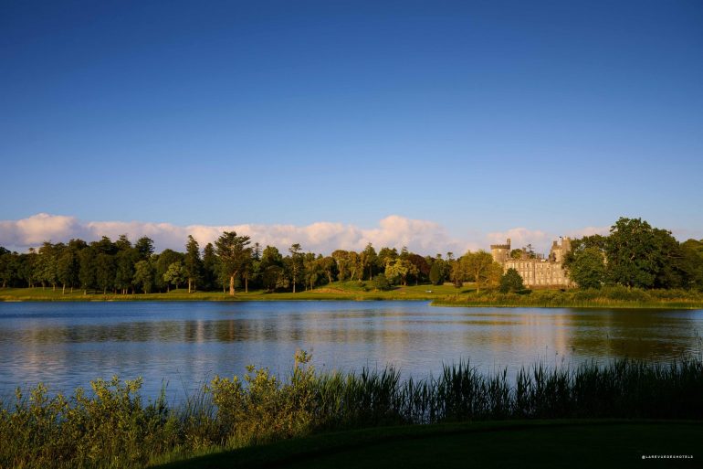 Wide lake scene with a grassy bank, a line of trees, and a stone castle on the far shore under a clear blue sky.
