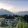 Aerial view of an Alpine resort with triangular A-frame cabins among pine trees and a large central lodge, mountain peaks in the distance at sunset