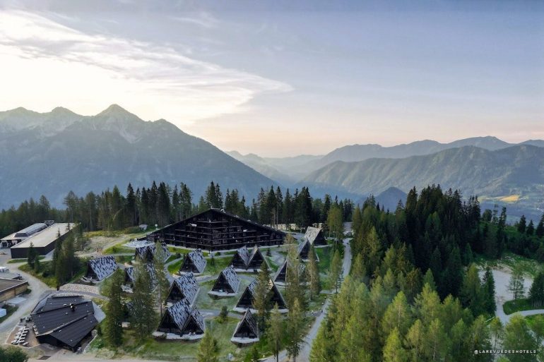 Aerial view of an Alpine resort with triangular A-frame cabins among pine trees and a large central lodge, mountain peaks in the distance at sunset