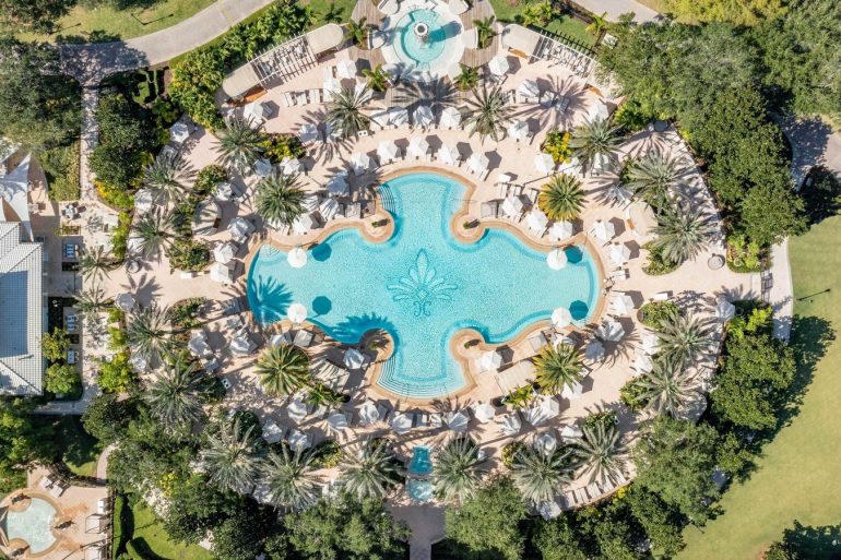 Aerial view of a resort pool area surrounded by palm trees, umbrellas, and lounge chairs. Top-down view of a turquoise pool with curved alcoves and sun loungers at a luxury resort. Resort pool complex from above with palm-lined deck, white umbrellas, and sun loungers around a blue pool. n aerial shot of tropical landscaping surrounding a large blue pool at a resort. erial image of a circular pool surrounded by palm trees and cabanas at a resort.