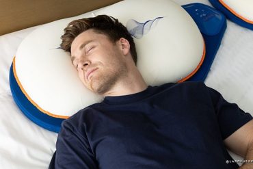 Man sleeping on a white neck pillow with blue edging on a bed far left.