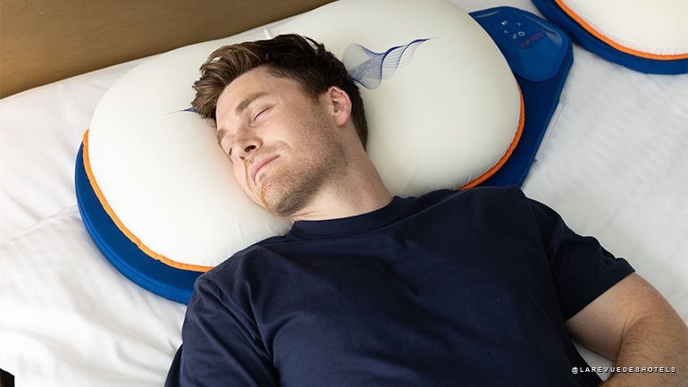 Man sleeping on a white neck pillow with blue edging on a bed far left.