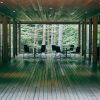 Wooden deck hallway with glass walls and black tables and chairs, opening to a forest view.