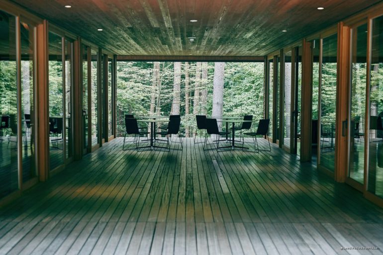 Wooden deck hallway with glass walls and black tables and chairs, opening to a forest view.