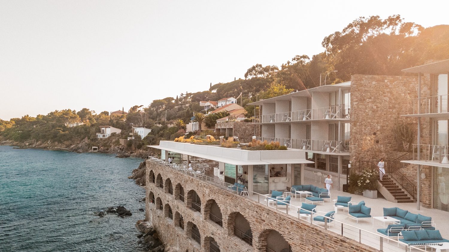 Resort terrace built into a rocky shoreline overlooking the sea with blue couches and white chairs.