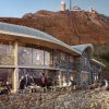 Stone hotel built into a mountain with a glass-walled dining area and outdoor terrace filled with guests.