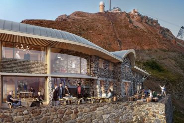 Stone hotel built into a mountain with a glass-walled dining area and outdoor terrace filled with guests.