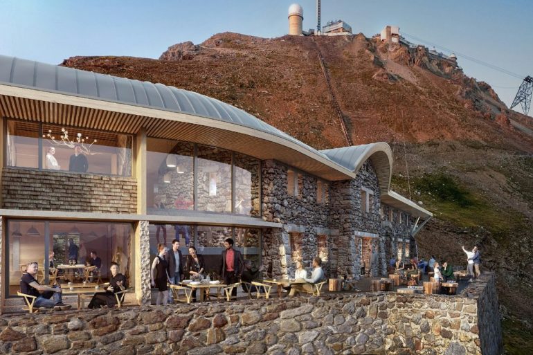 Stone hotel built into a mountain with a glass-walled dining area and outdoor terrace filled with guests.
