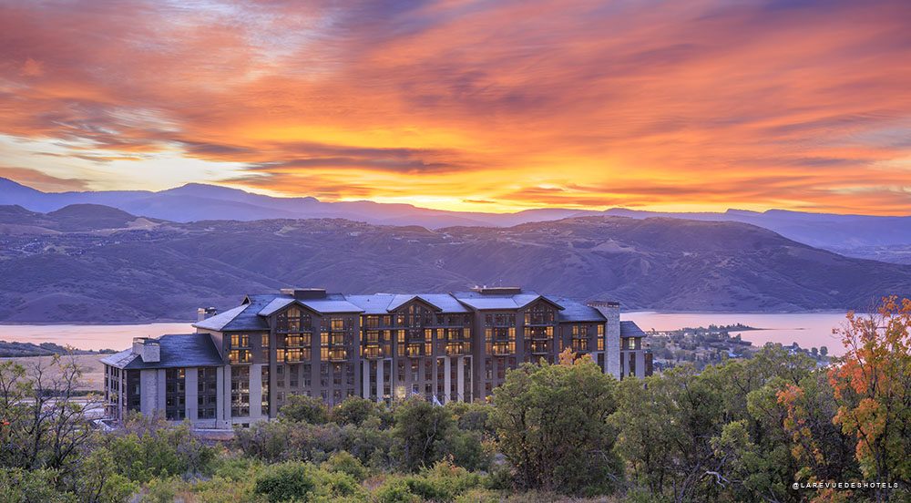 Luxurious resort hotel perched on hills with a sunset over a lake and purple mountains in the distance.
