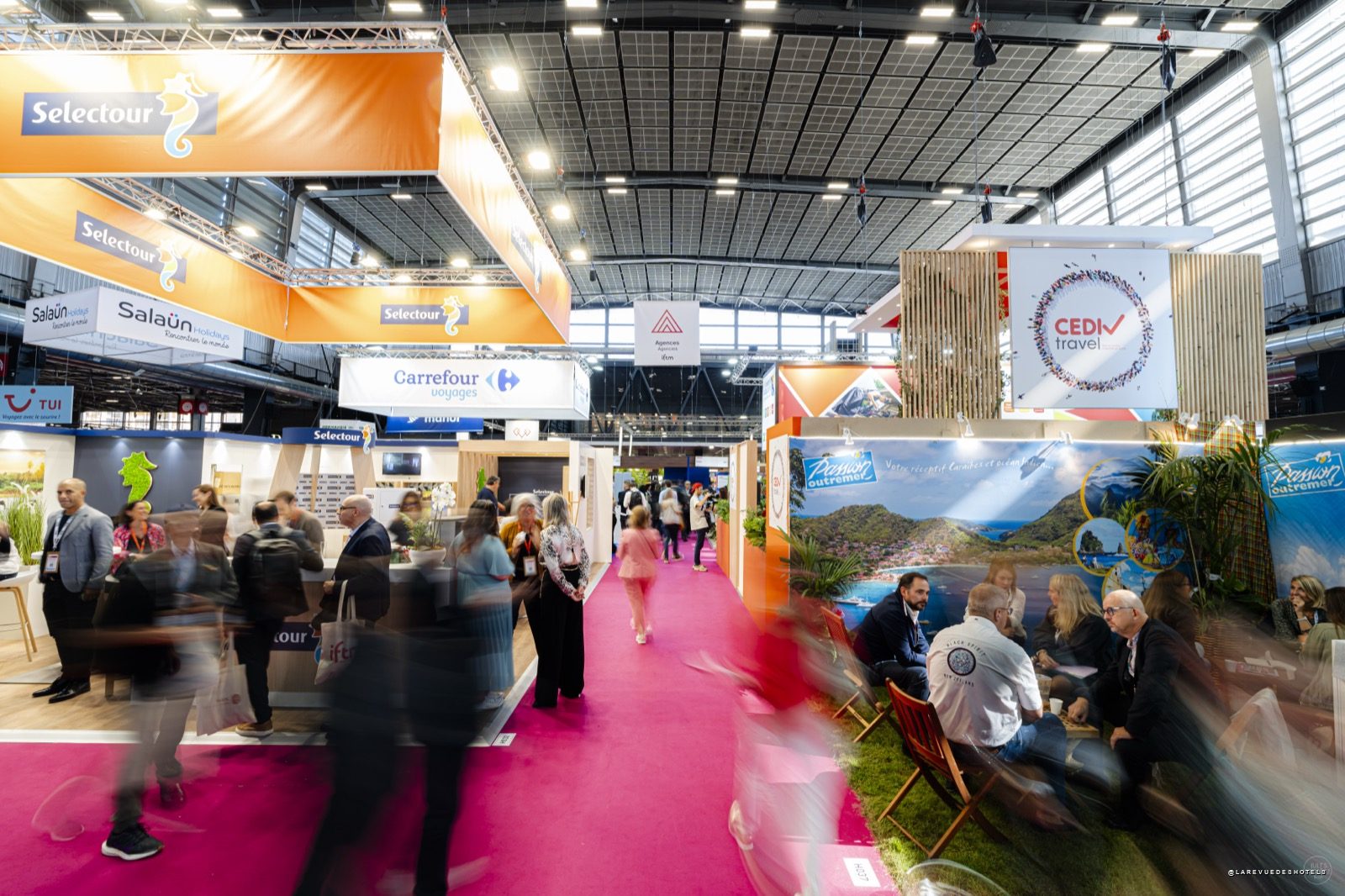 Busy exhibition hall with orange Selectour banners, pink carpet, and attendees visiting travel booths.