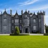 Gray stone castle with multiple towers sits on a manicured lawn under a blue sky, surrounded by trees at the rear.
