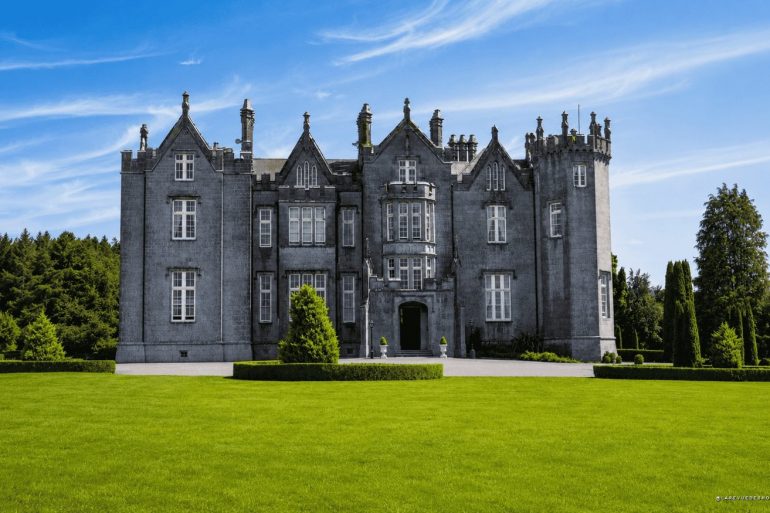 Gray stone castle with multiple towers sits on a manicured lawn under a blue sky, surrounded by trees at the rear.