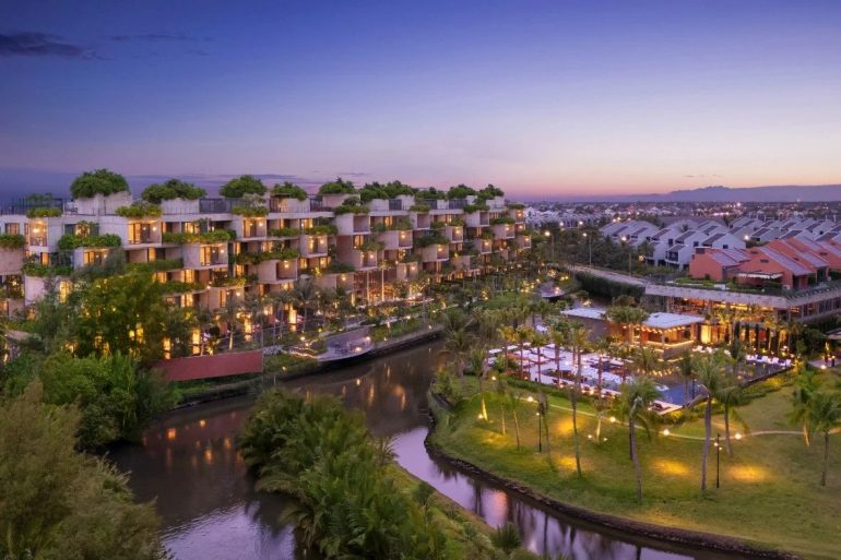 Resort complex at dusk with terraced buildings along a winding canal, palm trees and warm lights glowing at the terraces.