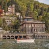 Elegant lakeside villa with arched stone quay and terrace, set on a forested hillside by calm waters; a small boat cruises by.