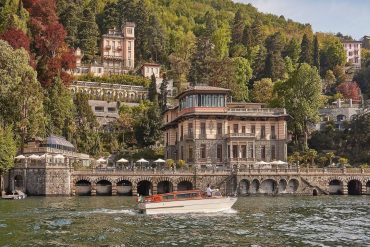 Elegant lakeside villa with arched stone quay and terrace, set on a forested hillside by calm waters; a small boat cruises by.