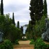 Bronze mermaid statues flank a gravel path in a lush garden, tall cypress trees framing a view of the sea in the distance.