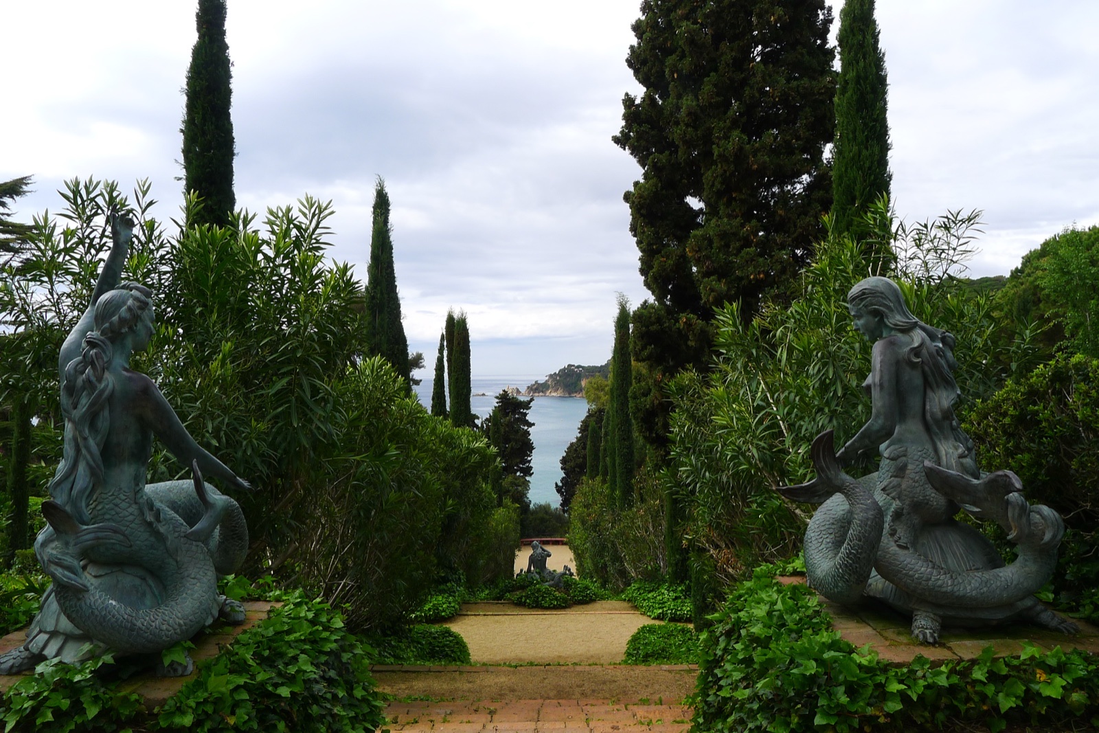 Bronze mermaid statues flank a gravel path in a lush garden, tall cypress trees framing a view of the sea in the distance.