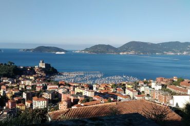 Coastal town with red-tiled roofs, a harbor filled with sailboats, and blue sea under a clear sky.