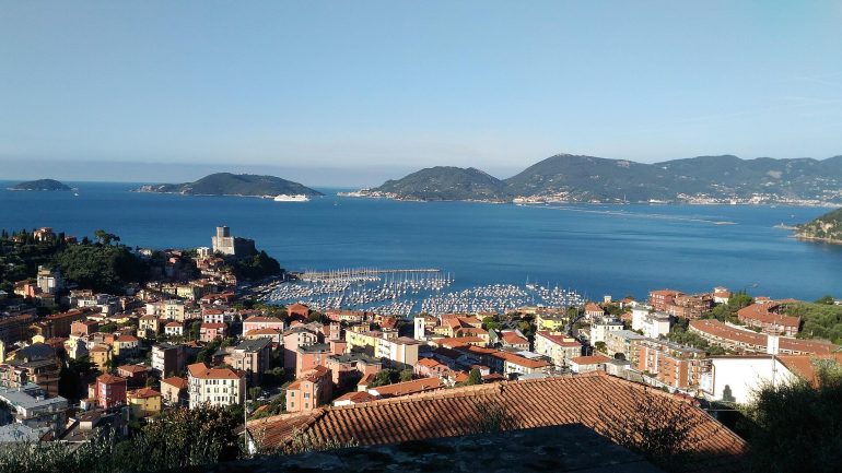 Coastal town with red-tiled roofs, a harbor filled with sailboats, and blue sea under a clear sky.