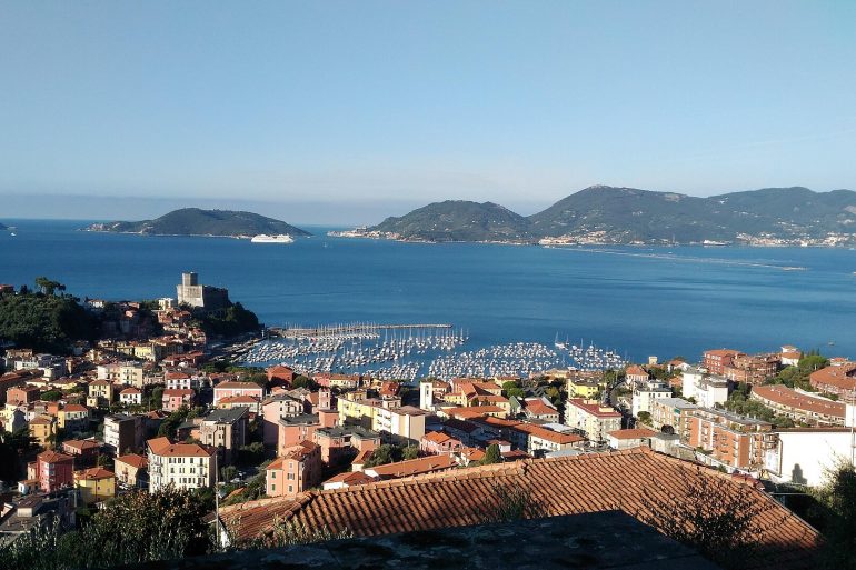 Coastal town with red-tiled roofs, a harbor filled with sailboats, and blue sea under a clear sky.