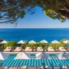 Resort pool area with white umbrellas and lounge chairs overlooking a blue sea, framed by tree branches above.