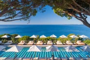 Resort pool area with white umbrellas and lounge chairs overlooking a blue sea, framed by tree branches above.