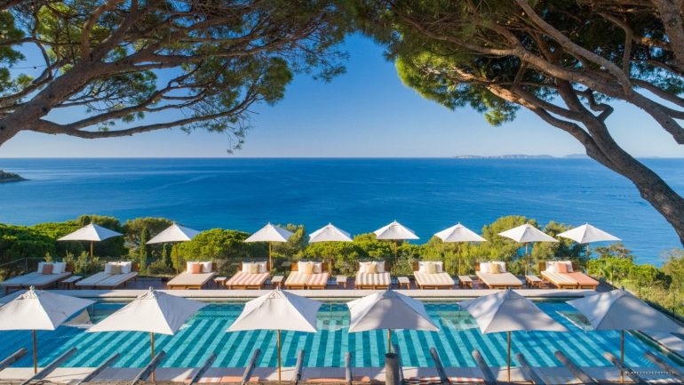 Resort pool area with white umbrellas and lounge chairs overlooking a blue sea, framed by tree branches above.
