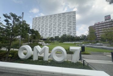 Large white 3D letters spell 'OMO7' in a landscaped plaza with a modern, irregular-patterned white building behind it.