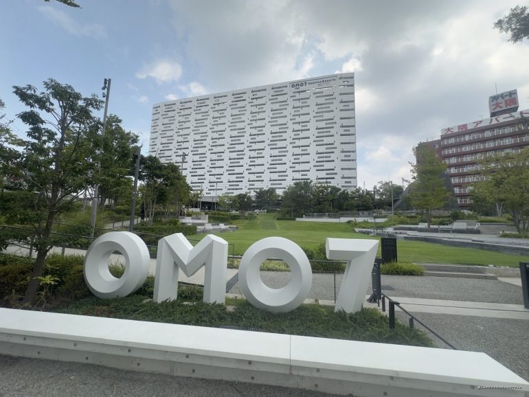 Large white 3D letters spell 'OMO7' in a landscaped plaza with a modern, irregular-patterned white building behind it.