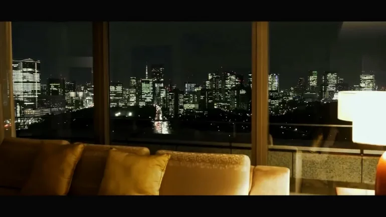 Nighttime city skyline seen through large glass windows from a living room with a beige sofa in the foreground.