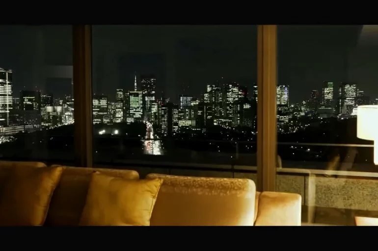 Nighttime city skyline seen through large glass windows from a living room with a beige sofa in the foreground.