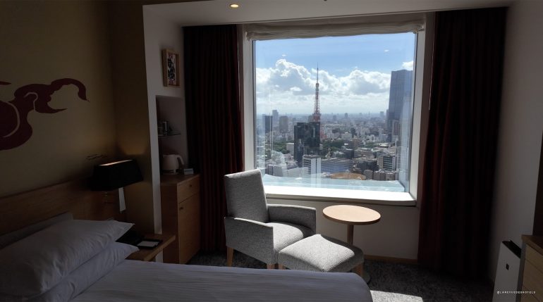Hotel room with a large window offering a city view, a gray armchair and small round table by the window.