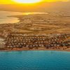 Aerial view of a coastal resort at sunset with white villas along a curved sandy shoreline and turquoise water nearby.