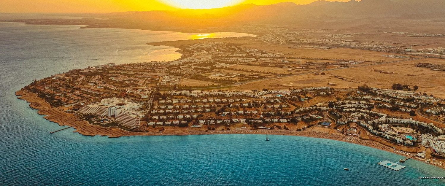 Aerial view of a coastal resort at sunset with white villas along a curved sandy shoreline and turquoise water nearby.