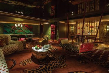 Opulent hotel lounge with leopard-print carpet, plush sofas, and a circular marble coffee table under warm lighting, plus hanging rope swings.