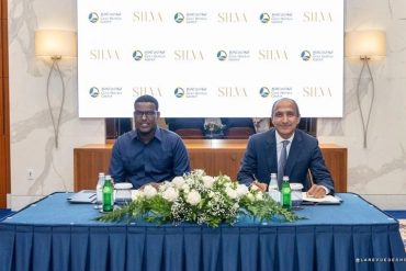 Two men in business attire sit at a table with a white flower centerpiece, against a backdrop featuring SILVA logos and hotel group branding.