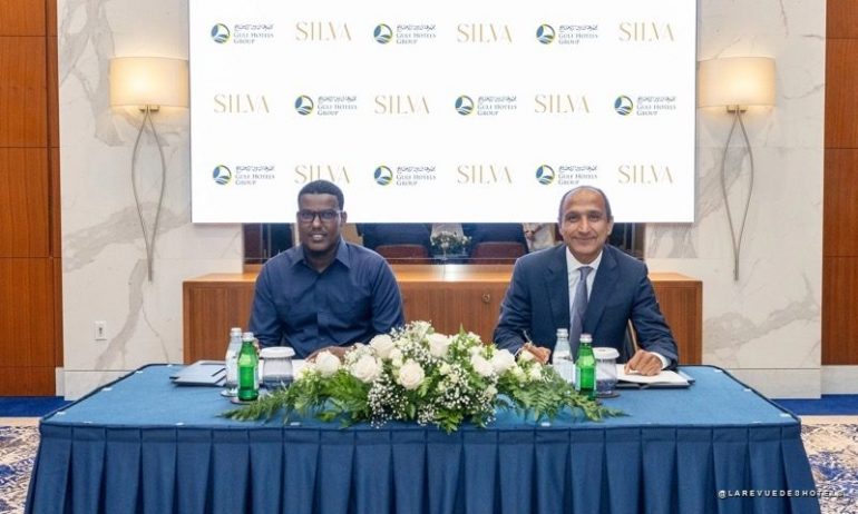 Two men in business attire sit at a table with a white flower centerpiece, against a backdrop featuring SILVA logos and hotel group branding.