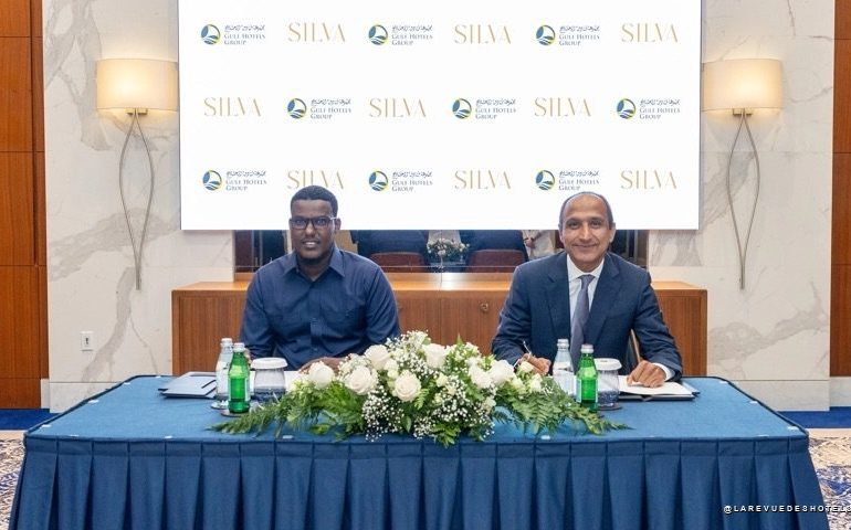 Two men in business attire sit at a table with a white flower centerpiece, against a backdrop featuring SILVA logos and hotel group branding.