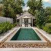 Rectangular swimming pool with a white pebble border and lush tropical plants, leading to a thatched-roof cabana with loungers.