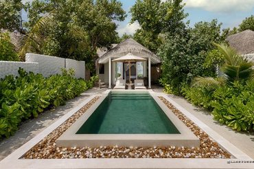 Rectangular swimming pool with a white pebble border and lush tropical plants, leading to a thatched-roof cabana with loungers.