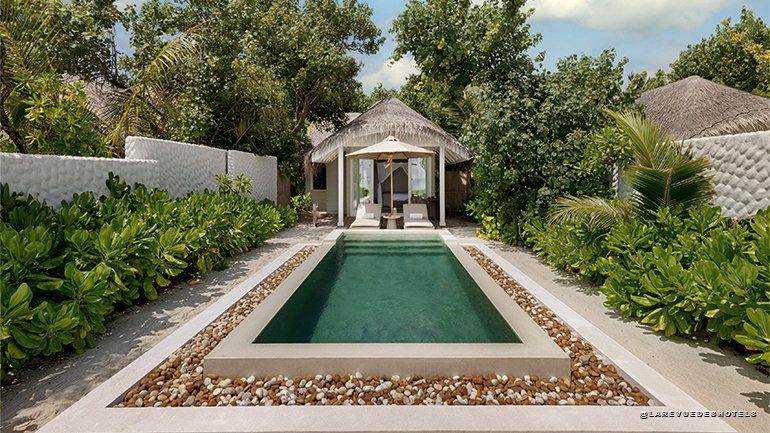 Rectangular swimming pool with a white pebble border and lush tropical plants, leading to a thatched-roof cabana with loungers.