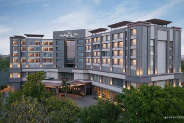 Exterior view of The Aarlis hotel: a modern, multi-wing building with warm-lit windows at dusk, surrounded by trees