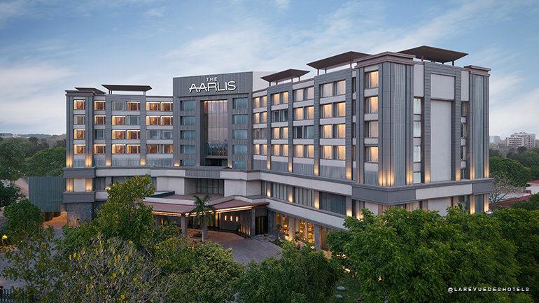 Exterior view of The Aarlis hotel: a modern, multi-wing building with warm-lit windows at dusk, surrounded by trees