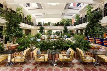 Bright hotel atrium with a central garden and yellow striped seating around marble-topped tables on a red-and-cream checkered floor.