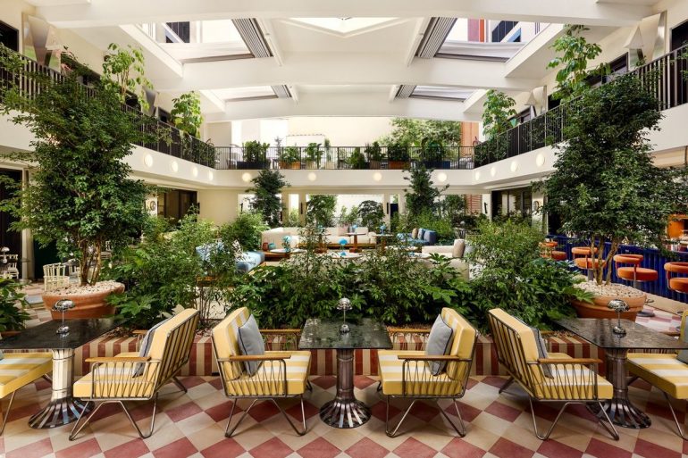 Bright hotel atrium with a central garden and yellow striped seating around marble-topped tables on a red-and-cream checkered floor.