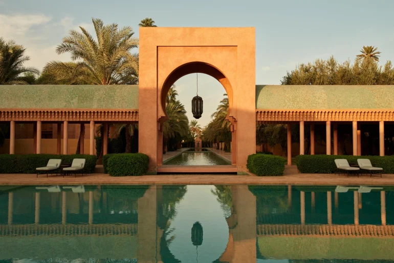 Piscine principale de l'Amanjena a Marrakech, arche mauresque rose et allee de palmiers au coucher du soleil