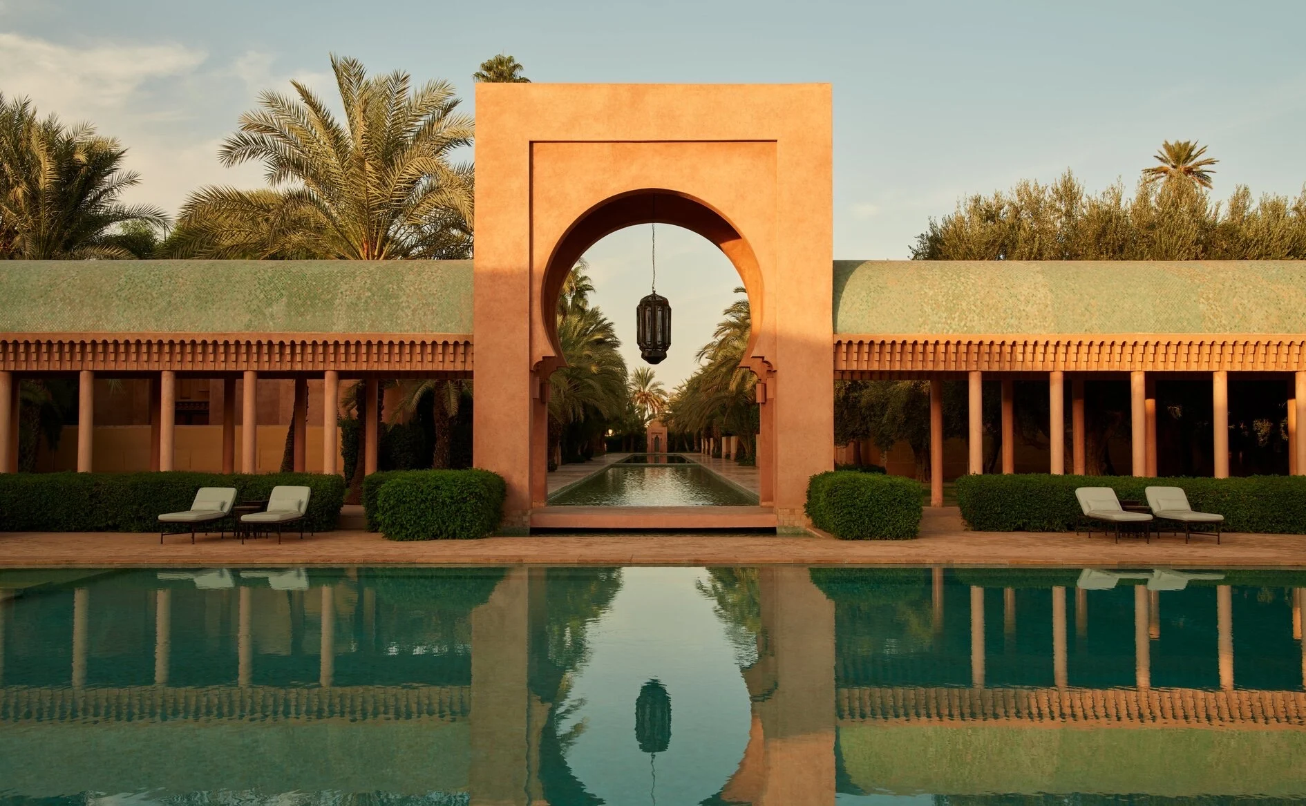 Piscine principale de l'Amanjena a Marrakech, arche mauresque rose et allee de palmiers au coucher du soleil